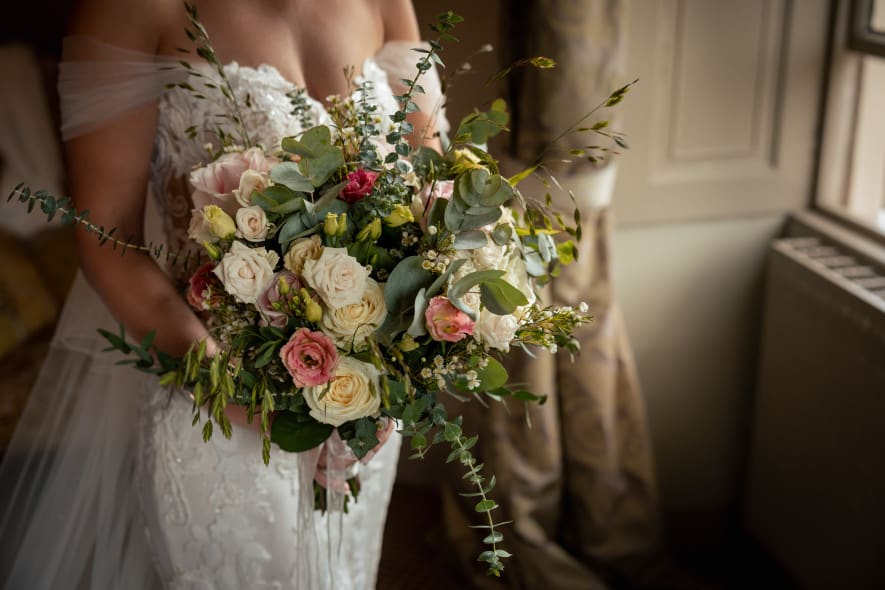 a bridge is wearing a white, lace off the should wedding dress, and is facing a window holding a large bouquet of flowers, with roses and eucalyptus