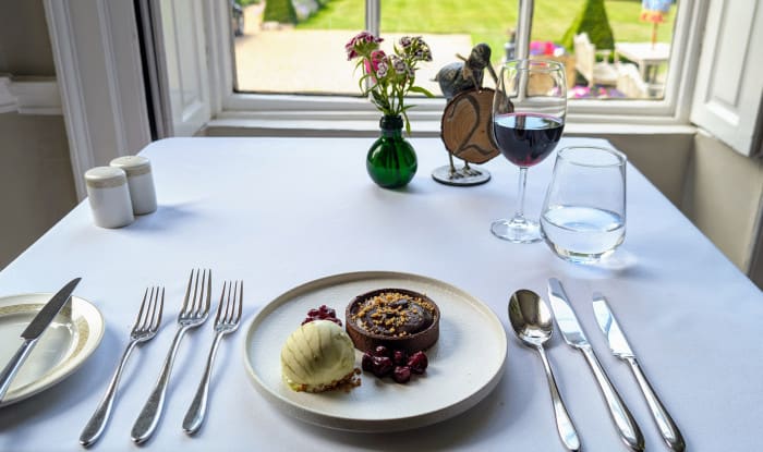 Chocolate tart with ice cream and cherries on a white plate, served at a window-side table with wine and a view of topiary garden.